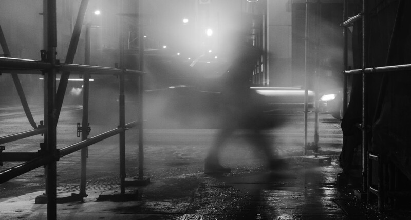 Blurred figure walking through wet urban street at night beneath street lights and building facades.