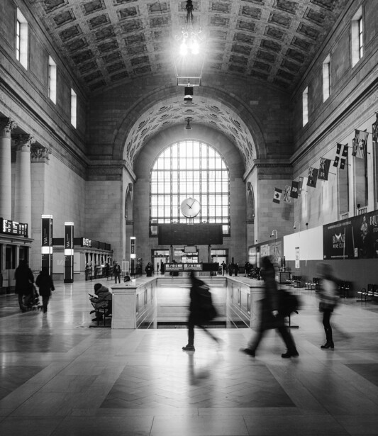 Black and white photograph of a grand interior hall with vaulted ceiling, large arched window, and blurred figures walking across polished floor.