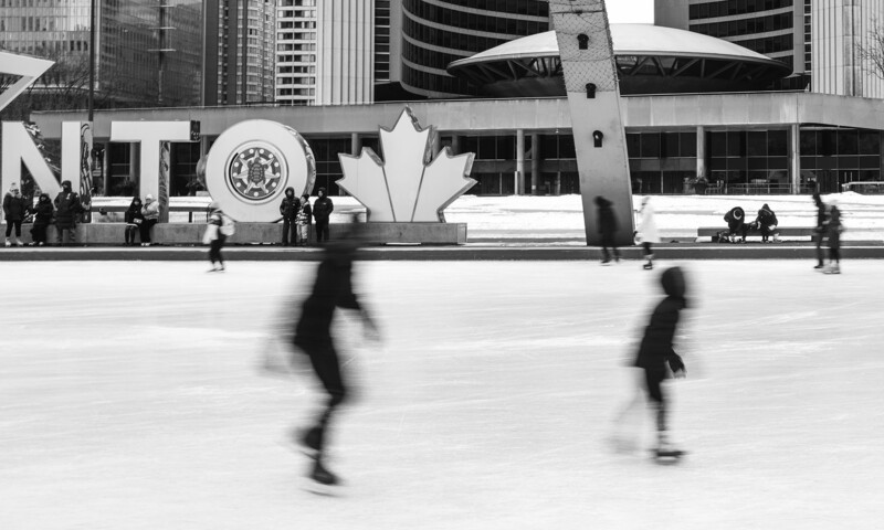 Blurred figures skating on an outdoor rink with modernist architecture and public sculptures in the background.