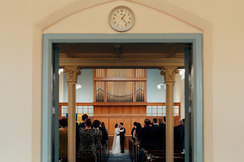 Wedding ceremony framed through a doorway with guests seated in pews and an ornate wooden altar visible.