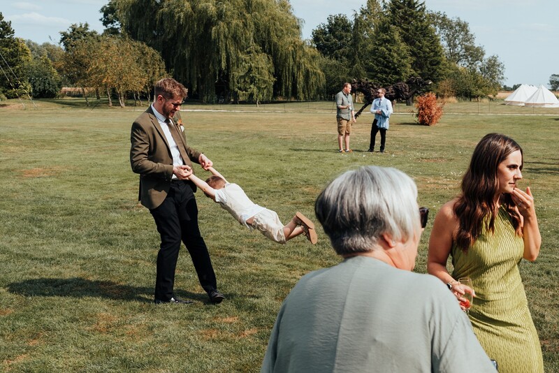 Man in brown blazer swings a child wrapped in white fabric while guests watch on a grassy field.