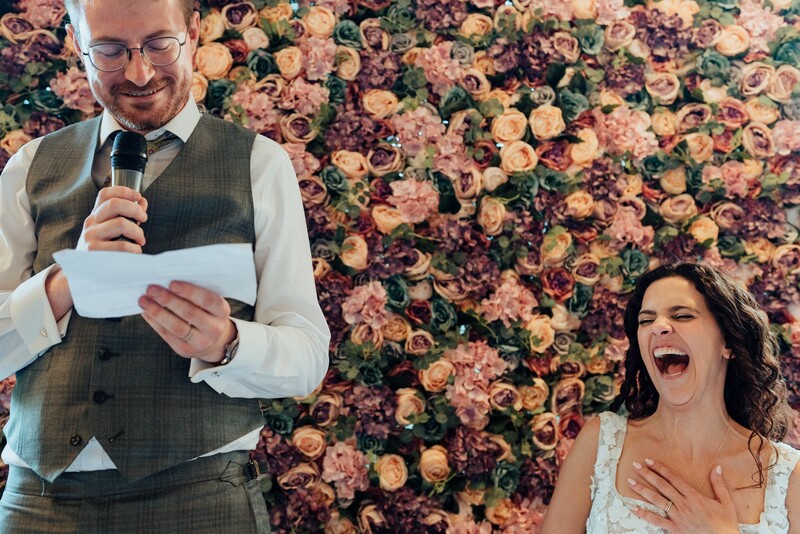 Man in vest and glasses speaking at microphone beside woman laughing in front of floral backdrop.