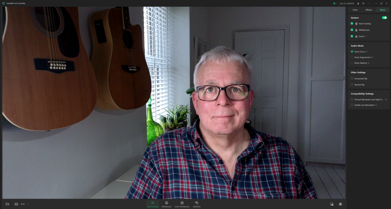 Screenshot of video call showing man wearing glasses and plaid shirt in home office with acoustic guitars on wall.