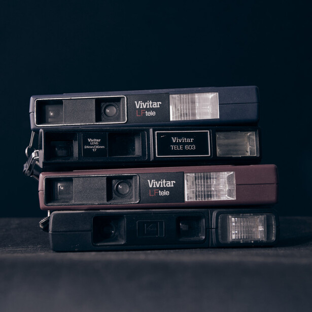 Stack of four vintage Vivitar rangefinder cameras with varying colored bodies in dark, neutral studio lighting.