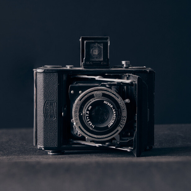 Vintage folding camera with bellows extended, black finish, and textured leather grip displayed against dark background.