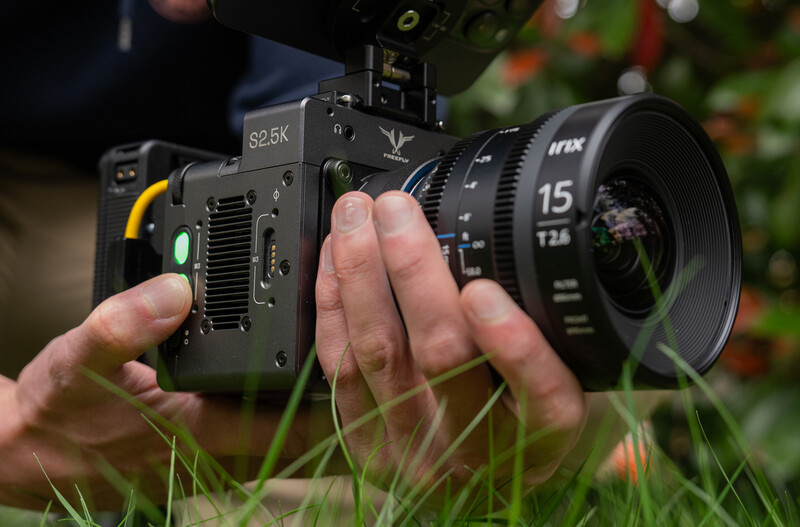 Hands holding a digital cinema camera with a 15mm lens attached, displayed outdoors with grass in foreground.