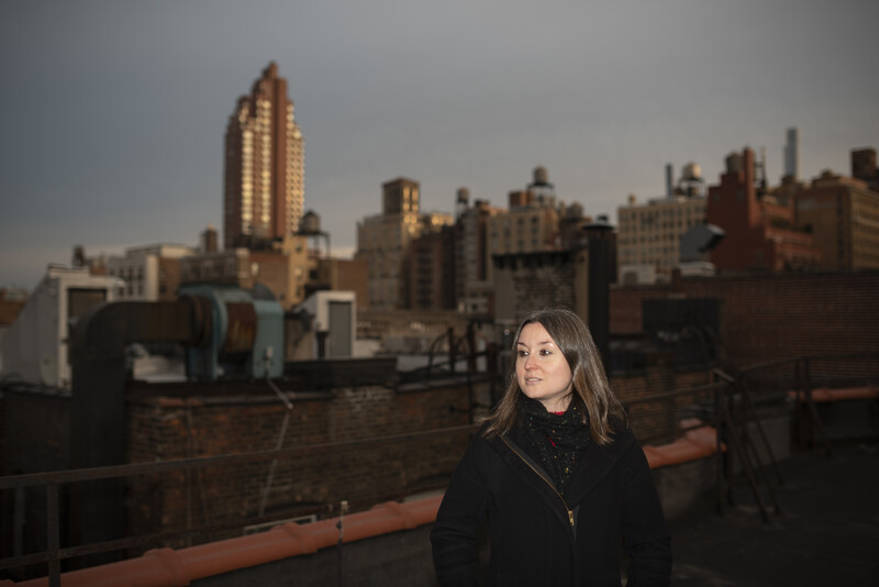 Woman in dark jacket looking over shoulder on rooftop with Manhattan skyline at golden hour.