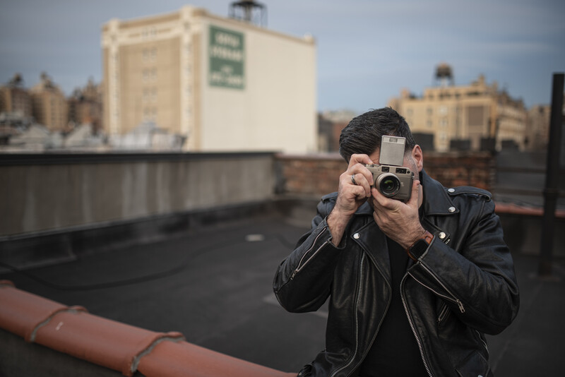 Photographer holding a rangefinder camera on a rooftop overlooking an urban skyline.