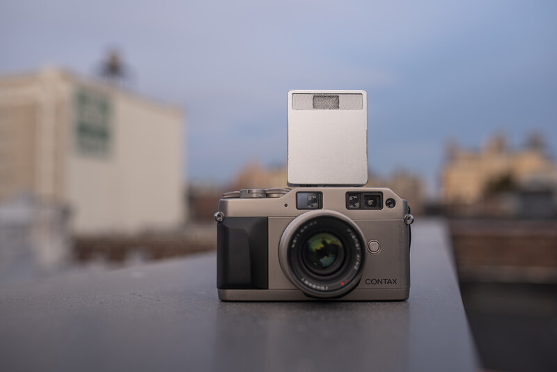 Contax rangefinder camera with external flash mounted on top, positioned on a surface with blurred urban buildings in the background.