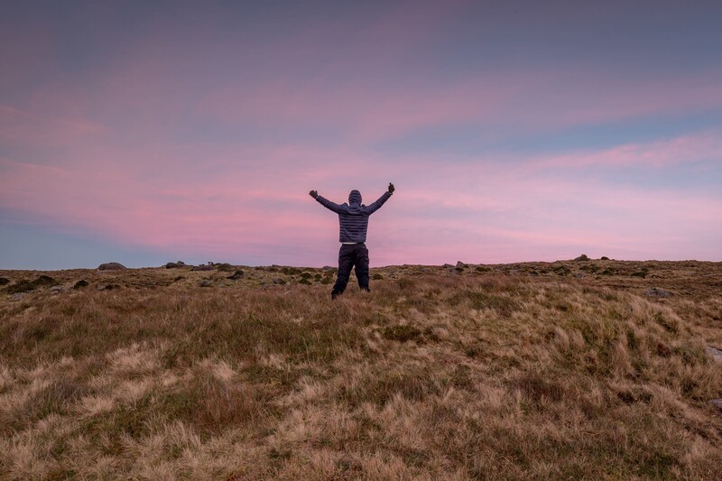 Person standing with arms raised in an open grassland under a pink and purple sunset sky.