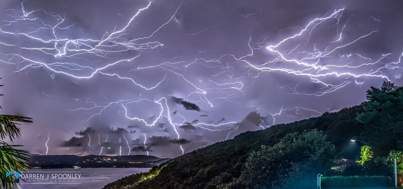 Intense lightning storm with branching bolts illuminating purple sky above a dark landscape with water and forested hills.