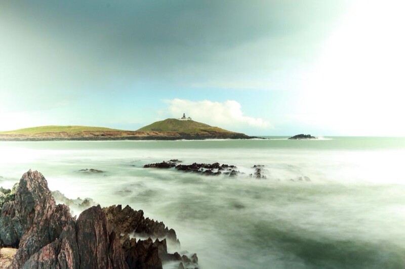 Rocky coastal rocks in foreground with long-exposure seascape, distant island with lighthouse, and soft green-tinted water.