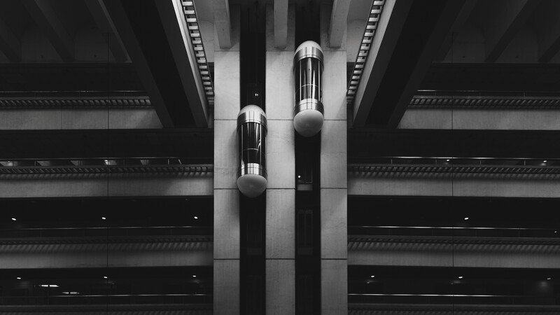 Two cylindrical elevator cars suspended vertically between concrete beams in a stark black and white architectural photograph.