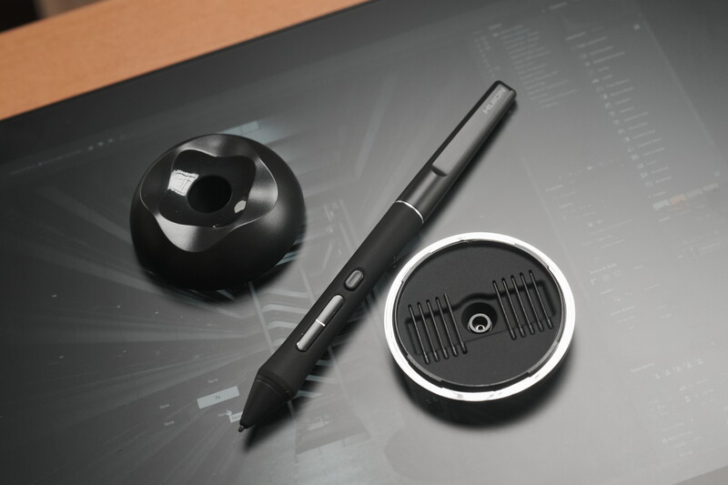 Three pieces of creative equipment arranged on a gray surface: a black spherical object, a stylus pen, and a circular device with a white rim.