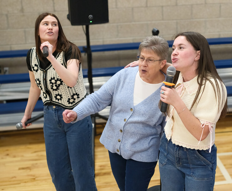 Three women speaking on a gymnasium floor with microphones during an indoor event.