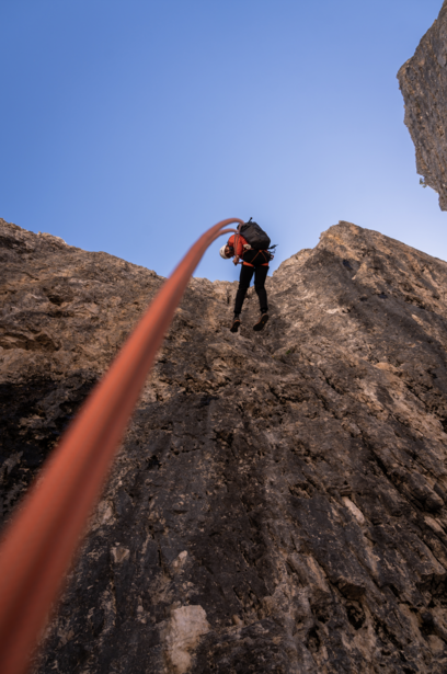 Rock climber ascending a steep natural rock face with a red rope against clear blue sky.