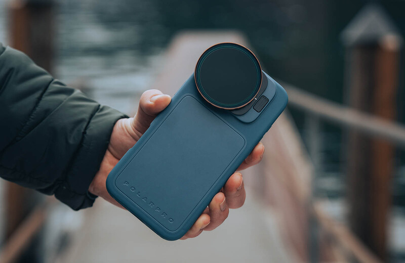 Hand holding a blue compact camera with a circular lens attachment in an outdoor setting.