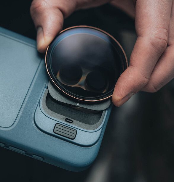 Hands holding a smartphone with a circular camera lens filter or attachment positioned on its lens.