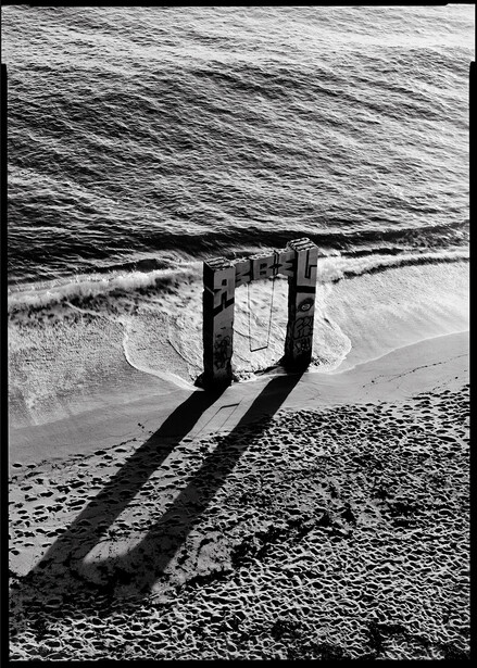 Wooden pier pilings casting long shadows across beach sand and shallow water in black and white.