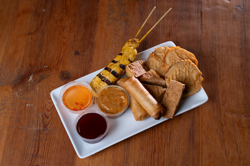 Overhead flatlay of a food styling composition with grilled protein skewers, bread, dipping sauces, and condiments on a white rectangular plate.
