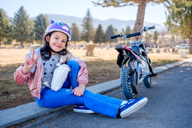 Child in blue helmet and protective gear sitting on pavement next to a blue children's bike, making a peace sign gesture.