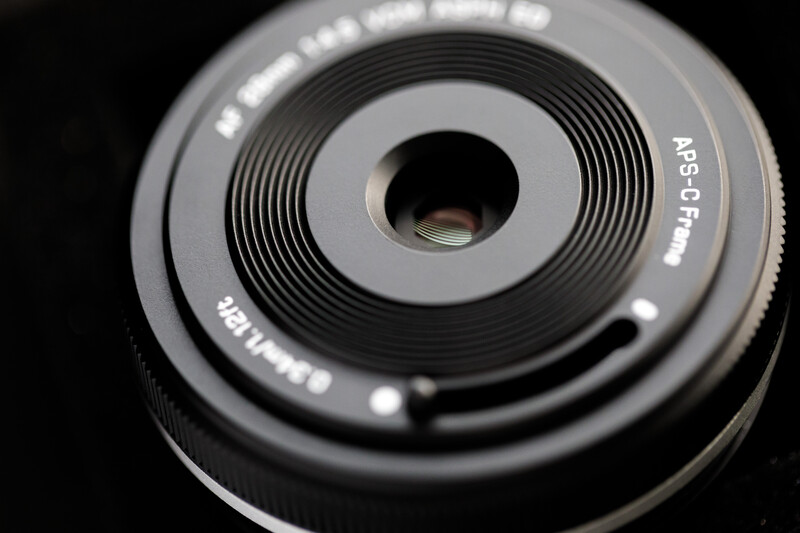 Close-up view of a compact camera lens with concentric aperture blades and central focusing element against black background.