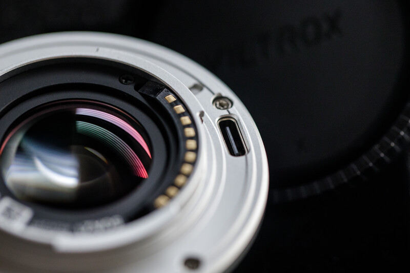 Close-up of a camera lens showing internal glass elements and gold-plated electrical contacts against a black background.