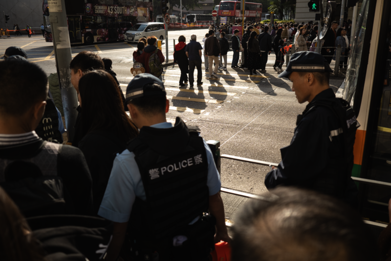 Police officers managing a crowd on a busy urban street with strong midday sunlight casting sharp shadows.