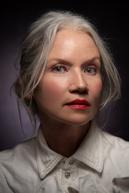 Portrait of a woman with gray hair wearing a light-colored collared shirt against a dark background.