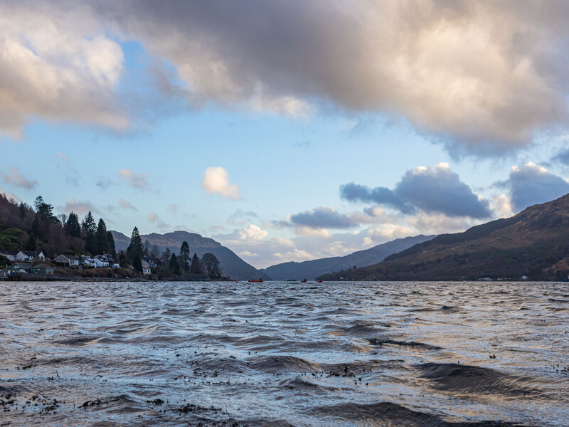 Mountainous lake landscape with coniferous trees along shoreline under dramatic cloudy sky.