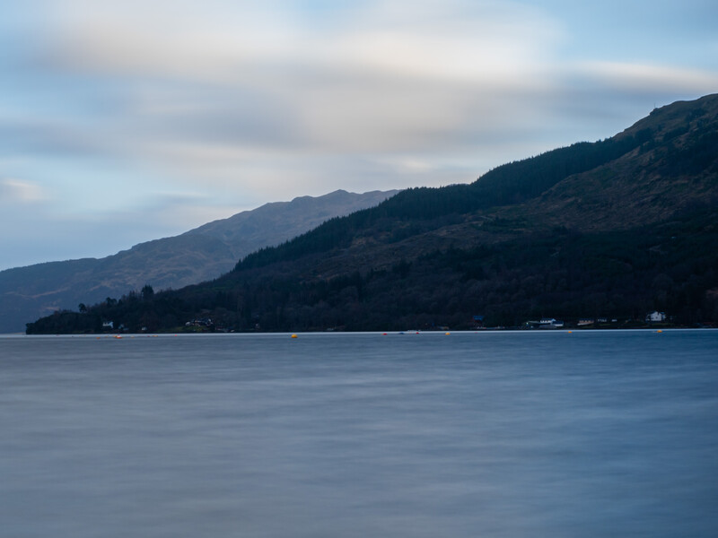 Calm lake with forested mountains in soft evening light under partly cloudy sky.