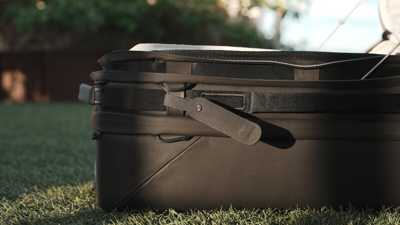 Black camera equipment bag sitting on grass with shoulder strap and handle visible in natural daylight.