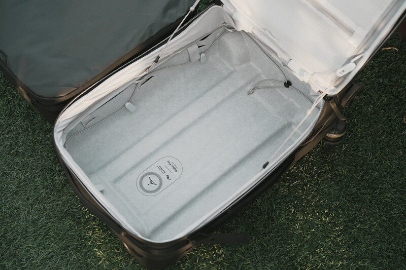 Open hard-shell suitcase with gray fabric interior and packing straps on green textured surface.