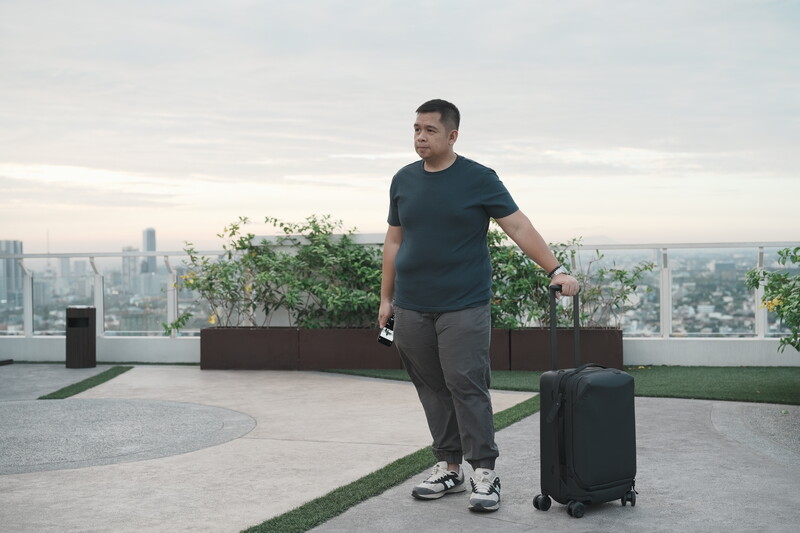 Man in dark t-shirt standing on rooftop with luggage overlooking city skyline at dusk.