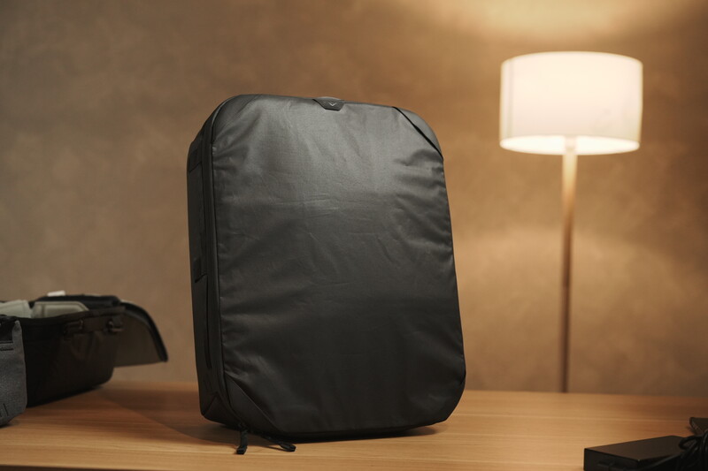 Black camera backpack displayed on wooden desk with warm studio lighting and floor lamp in background.