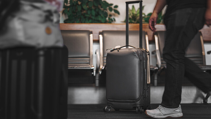 Black carry-on suitcase with extended handle in an airport terminal, with travelers and luggage visible in the background.