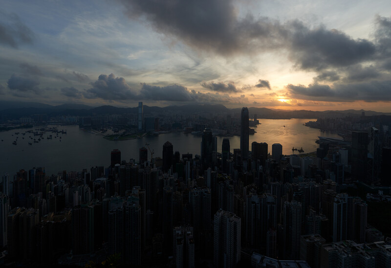 Dense urban skyline silhouetted against a golden sunset with dramatic storm clouds overhead and water visible in the foreground.