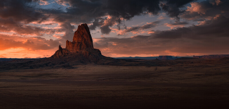 Dramatic desert landscape at sunset with a prominent red rock formation silhouetted against an orange and purple sky.
