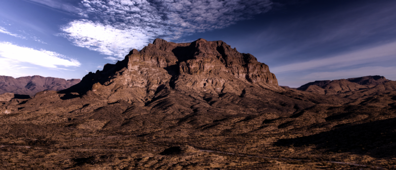 Rocky desert mountain with jagged peaks rising above arid, textured terrain under dramatic cloudy sky.
