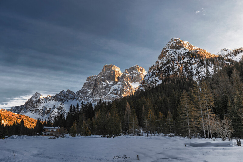 Snow-covered alpine lake surrounded by coniferous forest and dramatic rocky peaks bathed in golden sunlight.