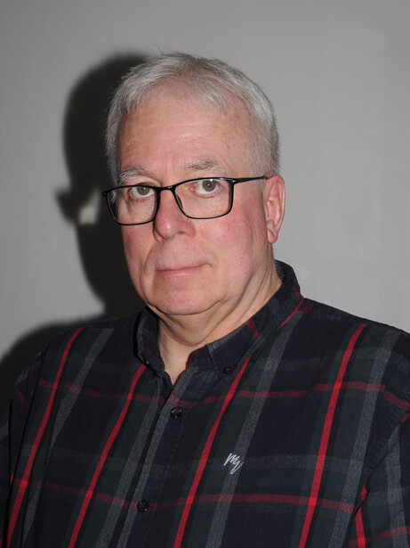 Middle-aged man with gray hair and glasses wearing a dark plaid shirt with red stripes against a neutral gray background.