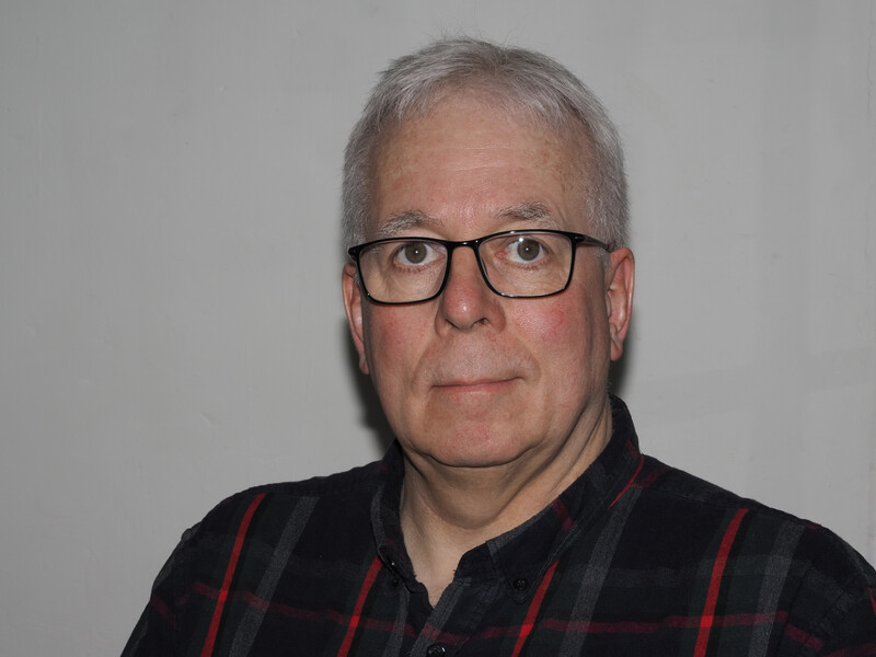 Middle-aged man with gray hair and glasses wearing a black and red plaid shirt against a neutral gray background.