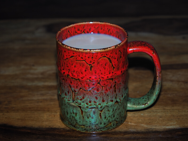 Ceramic mug with red crackle glaze on upper half and green weathered patina on lower half, sitting on wooden surface.