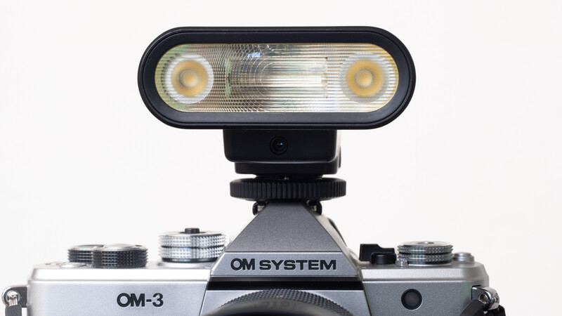 OM-3 camera with mounted flash unit featuring dual light sources and rounded design.