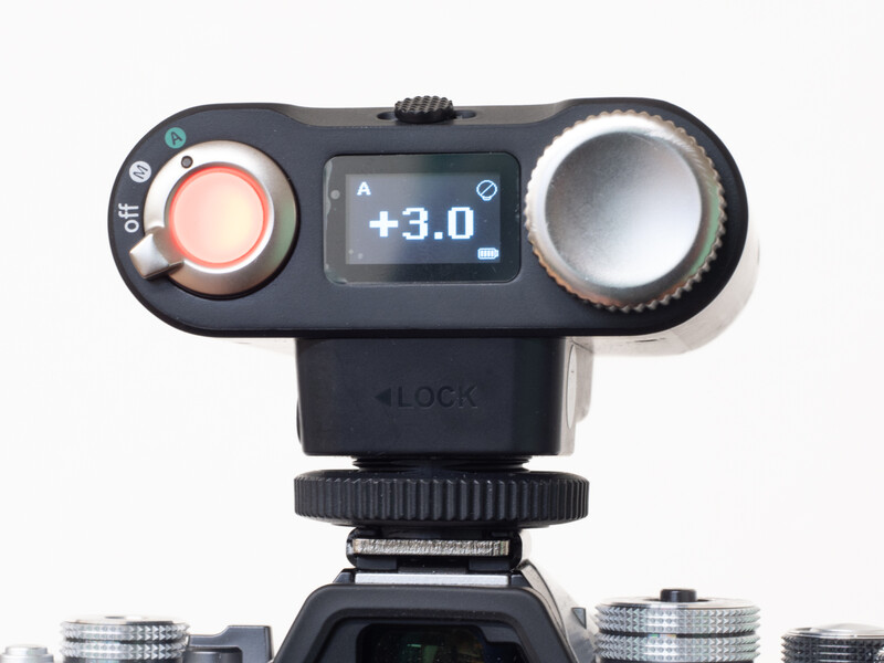 Top-down view of a camera-mounted exposure meter with dual dials and LCD display showing +3.0 aperture reading.