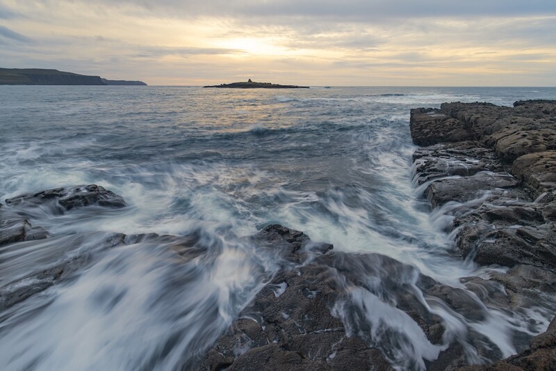 Waves crash over rocky coastline at dusk with distant headland visible across calm waters.