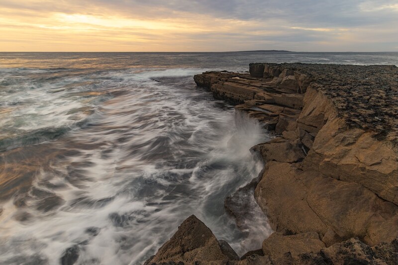 Waves crash against a rocky cliff formation as sunset light illuminates the layered stone and turbulent ocean.