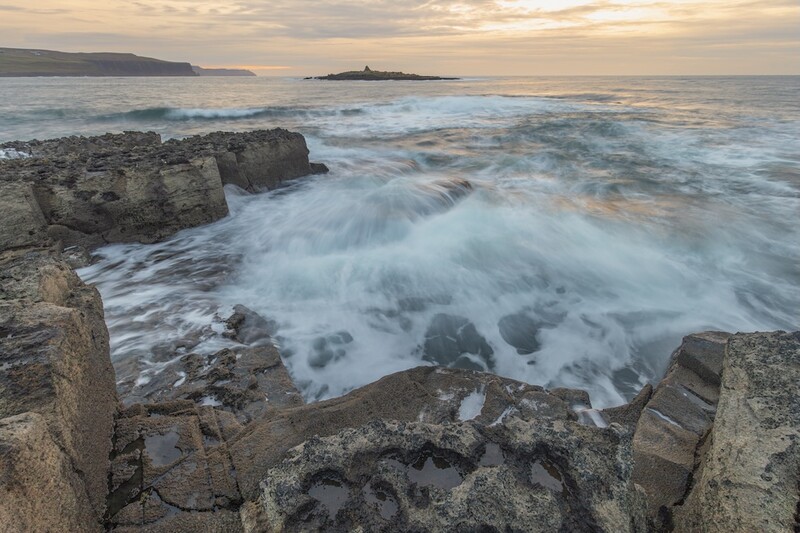 Seascape with foamy waves crashing over rocky foreground at golden hour, distant headland and island visible.