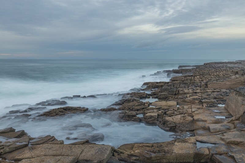 Rocky coastline with layered stone formations and long-exposure waves at dusk.