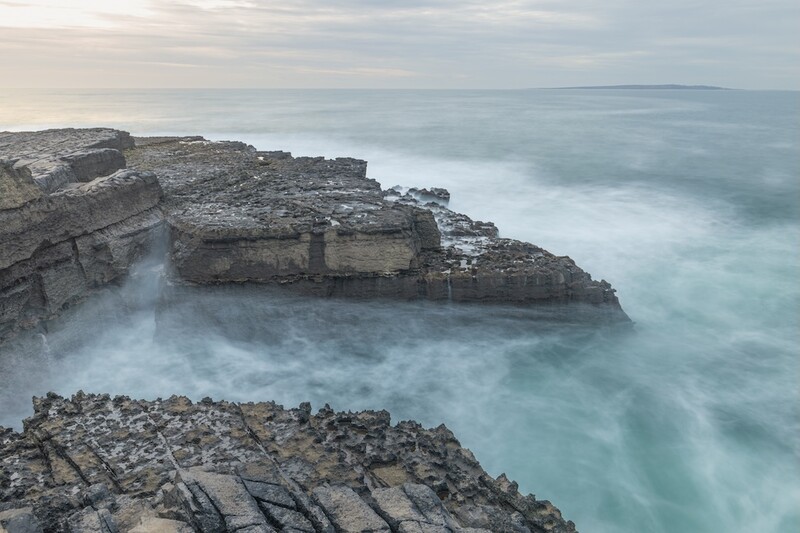 Layered rocky coastal cliffs with waves crashing below in turquoise water.